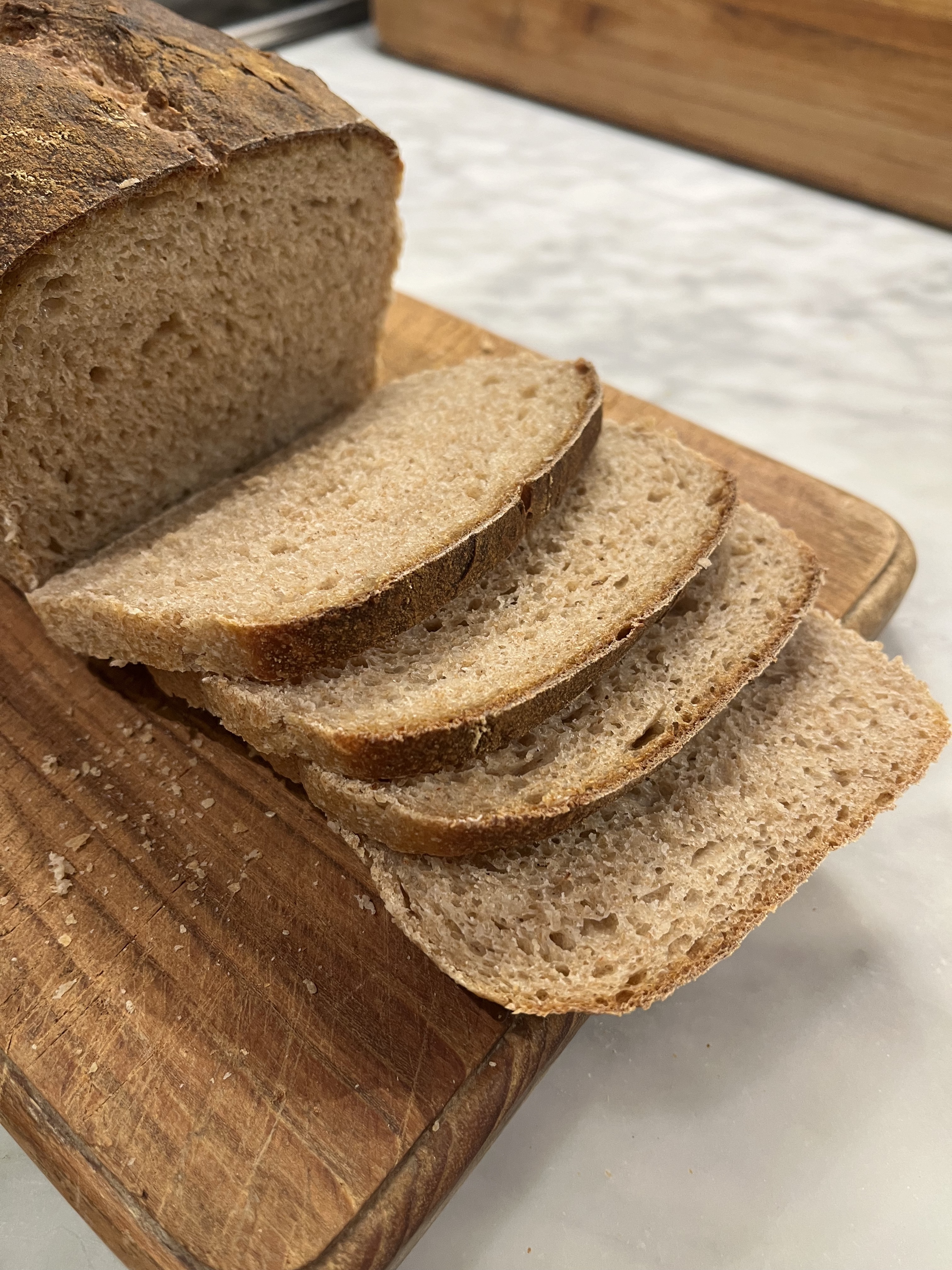 Photo of whole wheat sourdough sandwich bread partially sliced on a wood cutting board.