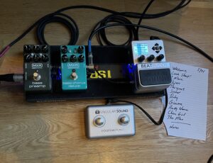 A small, bass pedalboard with preamp, chorus and Beat Buddy pedals on a wood floor with a hand written set list.